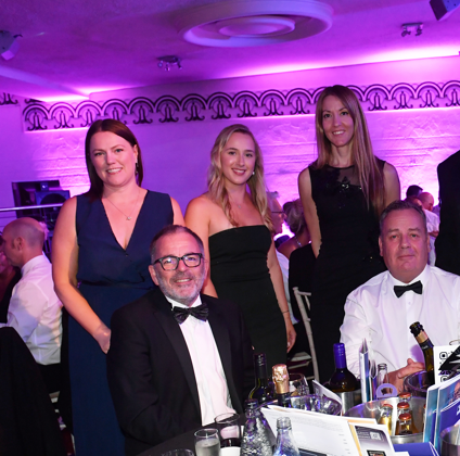 A group of five men in tuxedos and four women in dresses pose and smile around a table at the Constructing Excellence East Midlands Awards, with drinks and event programs on the table in the purple-lit room.