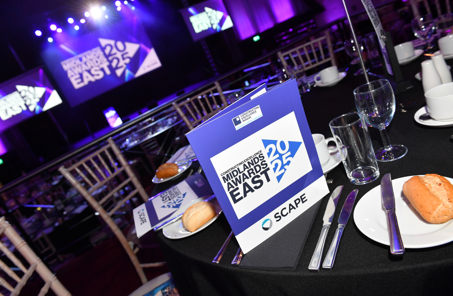 A close-up of a table set for the Constructing Excellence East Midlands Awards 2025, with a program, bread rolls, cutlery, and glasses. In the background, a stage and large display screens await the SME of the Year finalist, Waldeck.