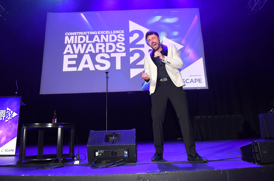 A man in a light jacket passionately speaks or sings on stage at the Midlands Awards East 2023, celebrating achievements like Constructing Excellence East Midlands’ SME of the Year, with stage lighting and an event banner in the background.