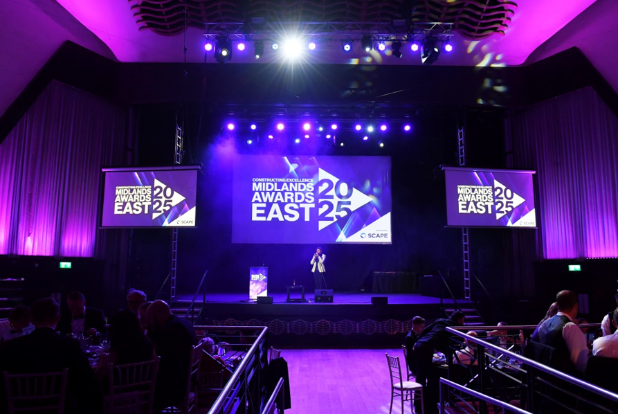 A presenter stands on stage at the Midlands Awards East 2025 event, with purple lighting, audience seating, and large screens displaying the SME of the Year award and Constructing Excellence East Midlands Awards.