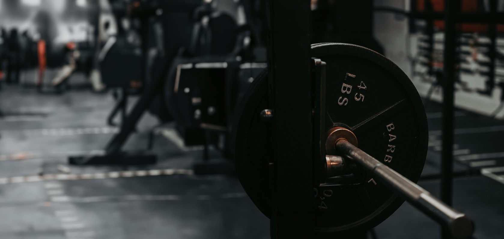 A gym with weightlifting equipment in focus, featuring a barbell loaded with 45-pound plates on a squat rack—perfect for Strong Girl Squad training—while other gym machines and weights blurred in the background.