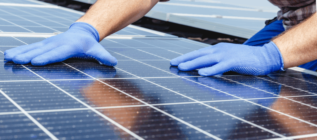 A person wearing blue protective gloves installs or adjusts solar panels outdoors, seeking to maximise efficiency and performance by carefully positioning both hands on the shiny surface of the panels.