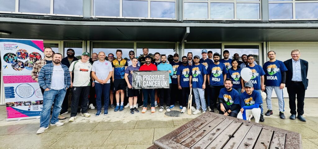 A large group of people, including the Waldeck nuclear team, pose outdoors—some holding cricket bats and a “Prostate Cancer UK” banner. Many wear blue India cricket shirts at this charity cricket match. A colorful display board sits to the left, with a table in front.