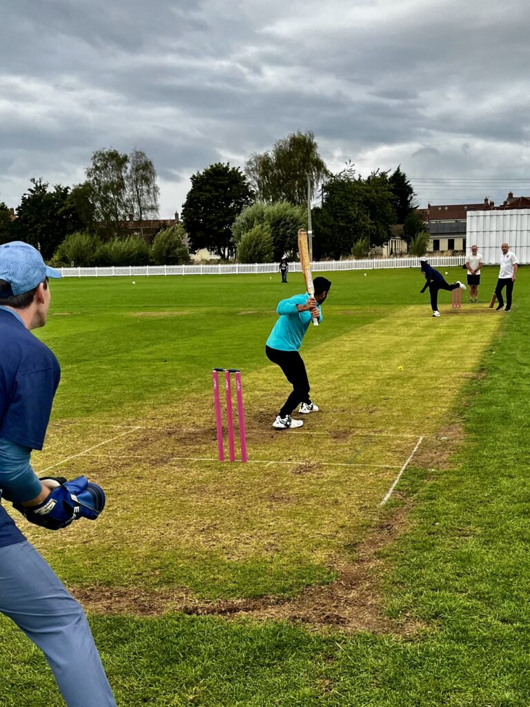 A charity cricket match unfolds on a grassy field; a batter in teal prepares to hit the ball, with a wicketkeeper behind and fielders in the background under a cloudy sky.