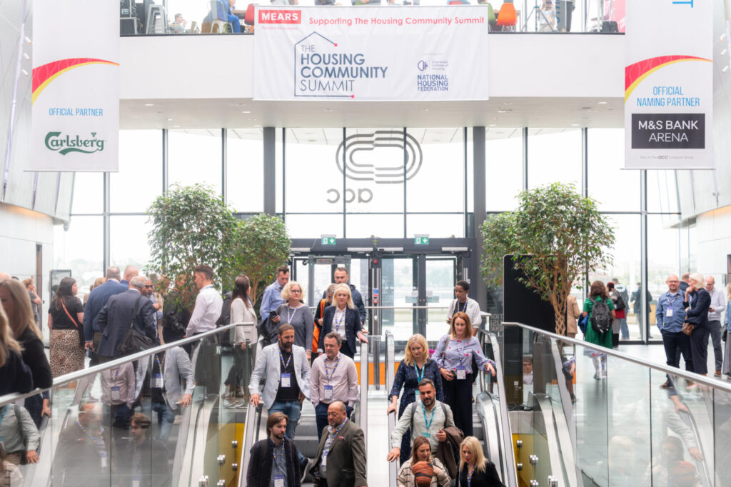 A diverse group of people, including industry leaders, attend the Housing Community Summit, moving through a bright, modern lobby with banners and large glass doors. Some are on escalators while others walk or talk in small groups.