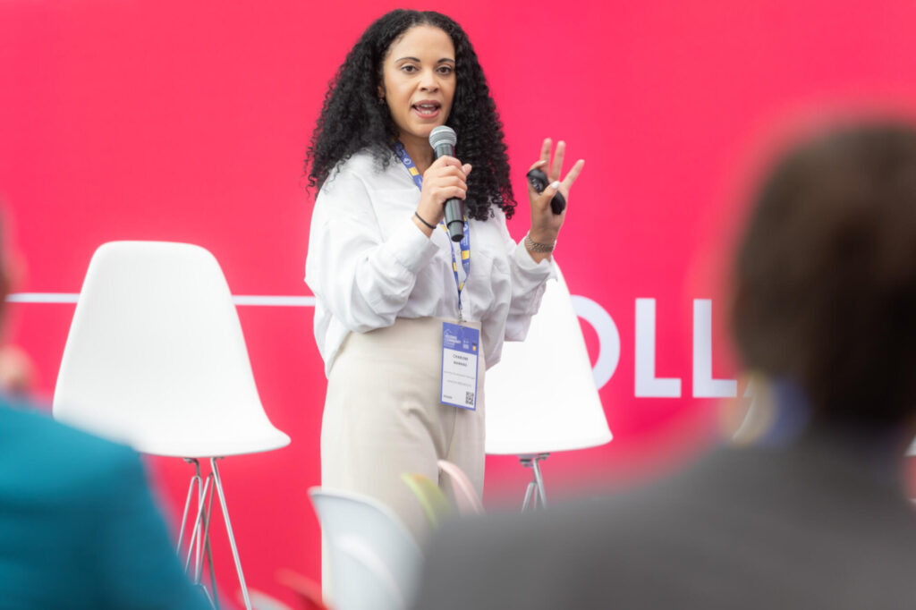 A woman with curly hair speaks into a microphone on stage at the Housing Community Summit, holding a small object, with empty white chairs and a bright pink background behind her. She wears a white blouse and beige pants.
