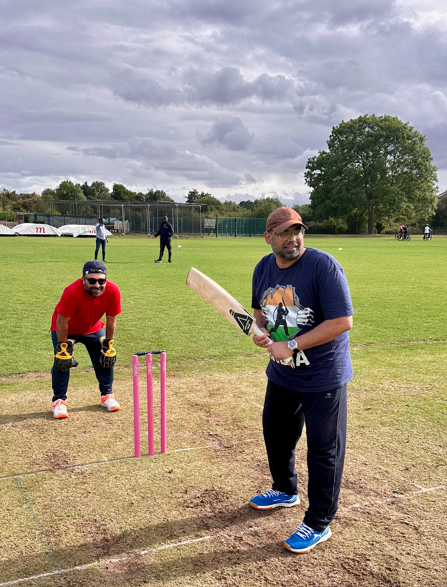Two men play cricket on a grassy field during a charity cricket match; one bats in a blue shirt, while the wicketkeeper in red crouches behind the stumps. Trees and a cloudy sky form the backdrop.