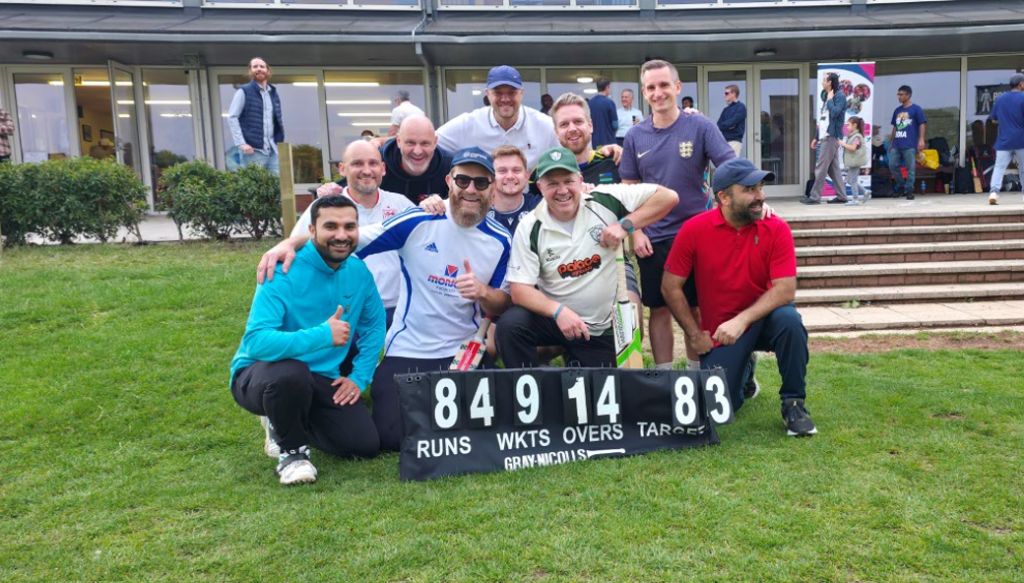 A group of ten men from the Nuclear Team pose and smile on a grassy field in front of a cricket scoreboard at the Waldeck Charity Cricket Match. Some wear cricket gear and hold bats, with a building visible in the background.