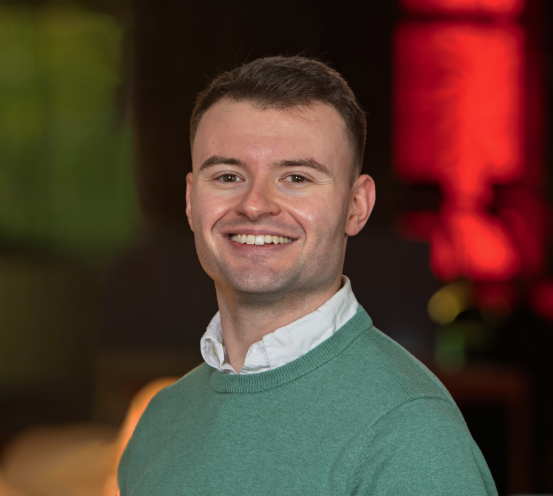 Josh Mahony, Senior Structural Technician, smiles at the camera. The young man with short brown hair wears a green sweater over a white collared shirt, set against a softly blurred background of warm red and green tones.