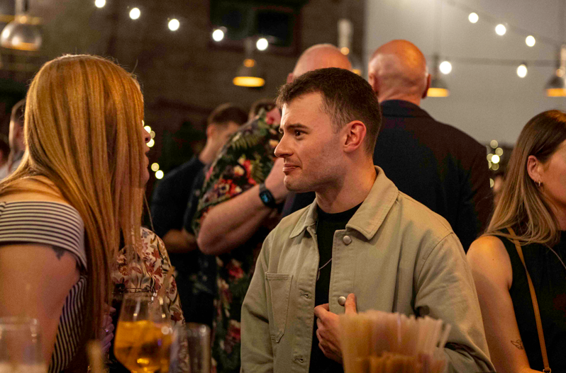 Senior Structural Technician Josh Mahony, in a light jacket, talks to a blonde woman at a lively indoor gathering, with people socializing and string lights overhead. Nearby, a woman with closed eyes sits holding a drink.