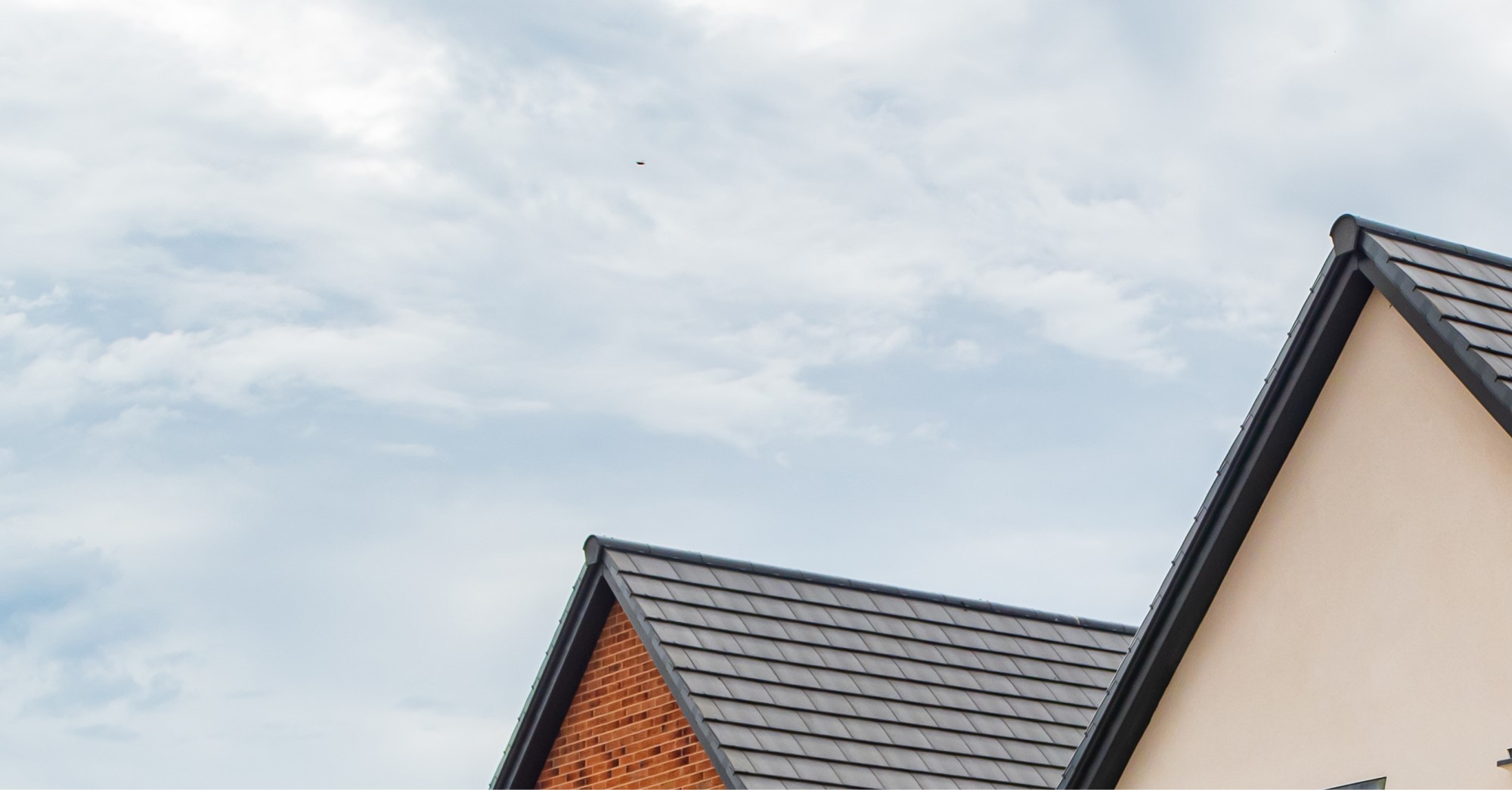 Two house rooftops with dark grey tiles and a cloudy sky above, showcasing housing regeneration in North West Leicestershire. One roof is attached to a cream-colored wall, and the other to a red brick wall.